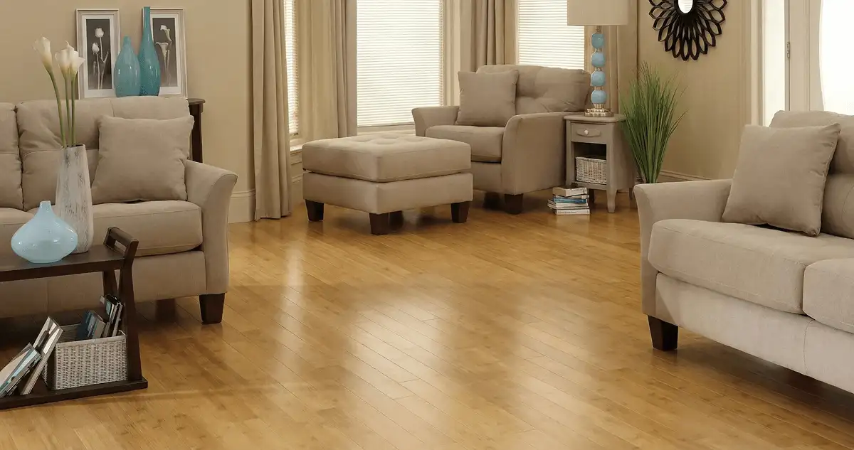 A living room with professionally installed bamboo flooring, beige sofas, a matching ottoman, a side table with a blue lamp, vases, framed pictures, and large windows with blinds and curtains in Little Rock, AR