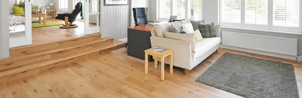 A modern living room with professionally installed laminate flooring, a beige sofa, a small side table, a gray rug, and large windows with white shutters in Little Rock, AR